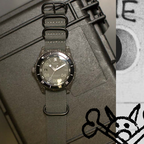 A dark grey Nixon watch with a fabric strap rests on a textured dark grey surface, with a partial black and white graphic visible on the right.