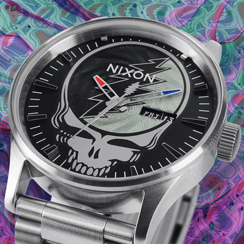 A close-up of a silver Nixon watch with a black face featuring the Grateful Dead "Steal Your Face" skull logo and a lightning bolt, set against a psychedelic background.