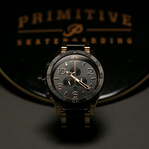 A Nixon watch with a dark grey face and rose gold numbers and hands is prominently displayed against a blurred background featuring "PRIMITIVE SKATEBOARDING" text.