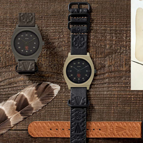 Two watches with dark faces and intricately embossed leather straps, one olive green and one tan, are displayed on a wooden surface next to a feather and an additional tan strap.