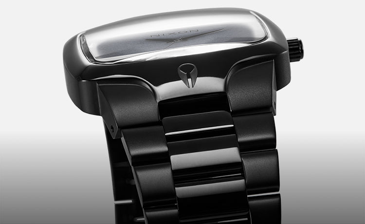 A close-up, angled view of a black Nixon watch with a rectangular face and a matching black metal band.