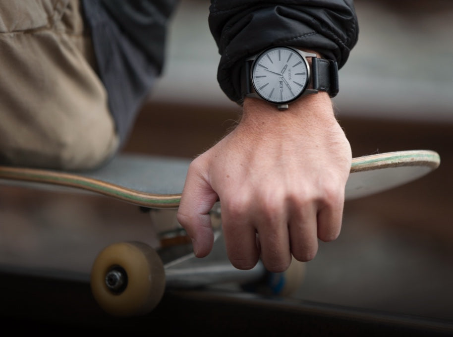 A skateboarder wears a Nixon Sentry Leather watch while sitting on his skateboard.